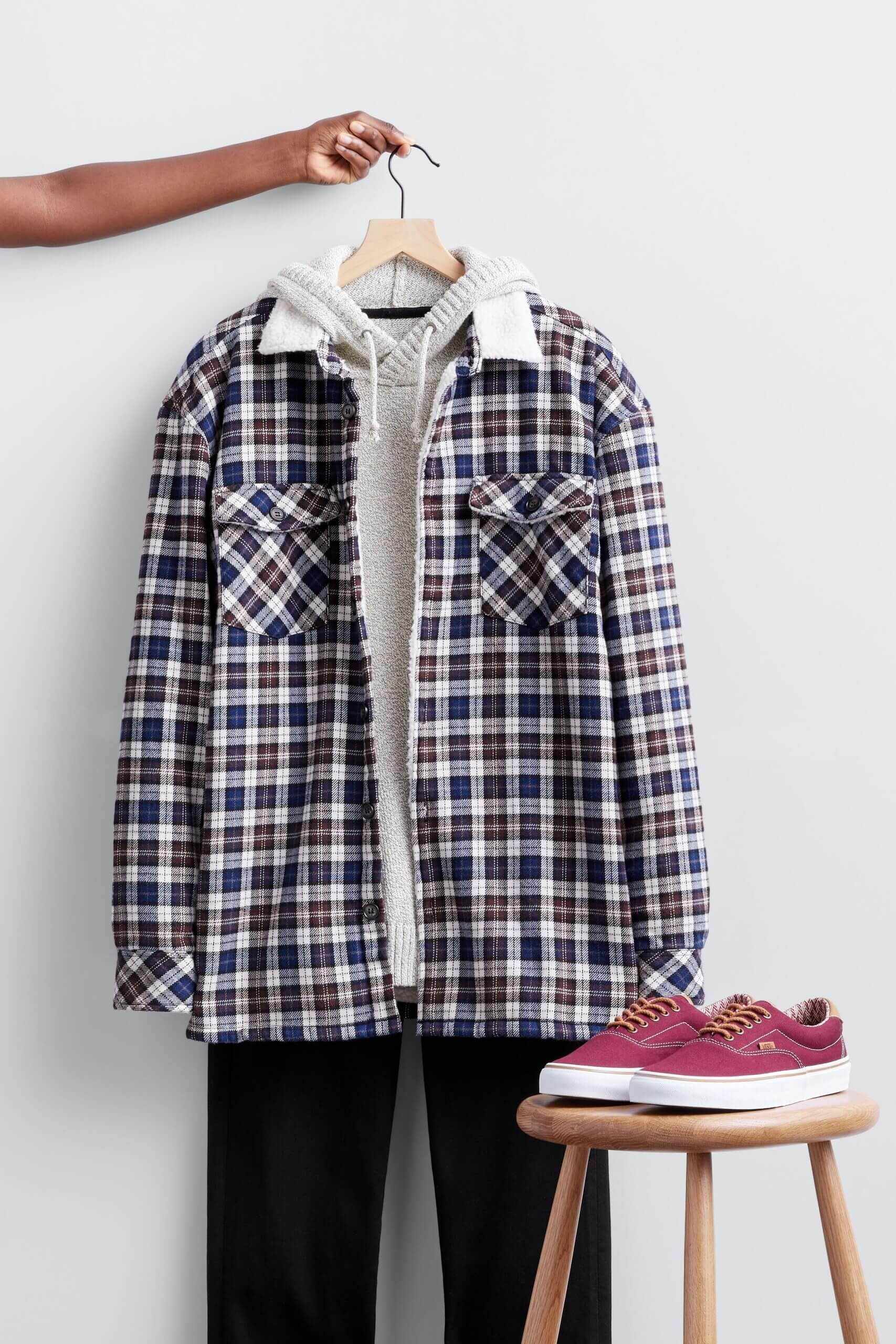 Stitch Fix Valentine's Day outfit for men featuring plaid shacket over hooded sweater and black jeans on hanger next to sneakers on stool.