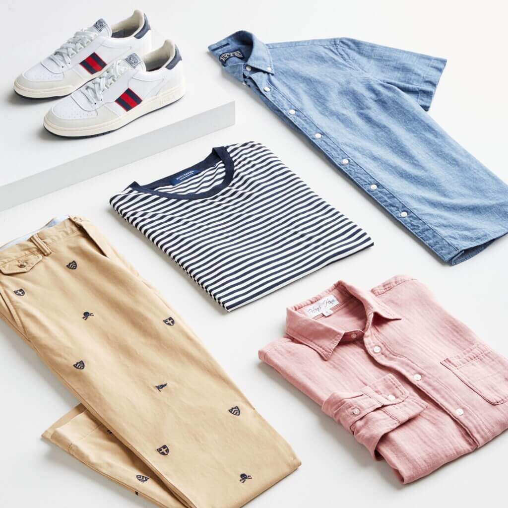 Stitch Fix men’s preppy style apparel featuring printed khaki pants, navy blue striped T-shirt, pink and blue button-down shirts and white lace-up sneakers.
