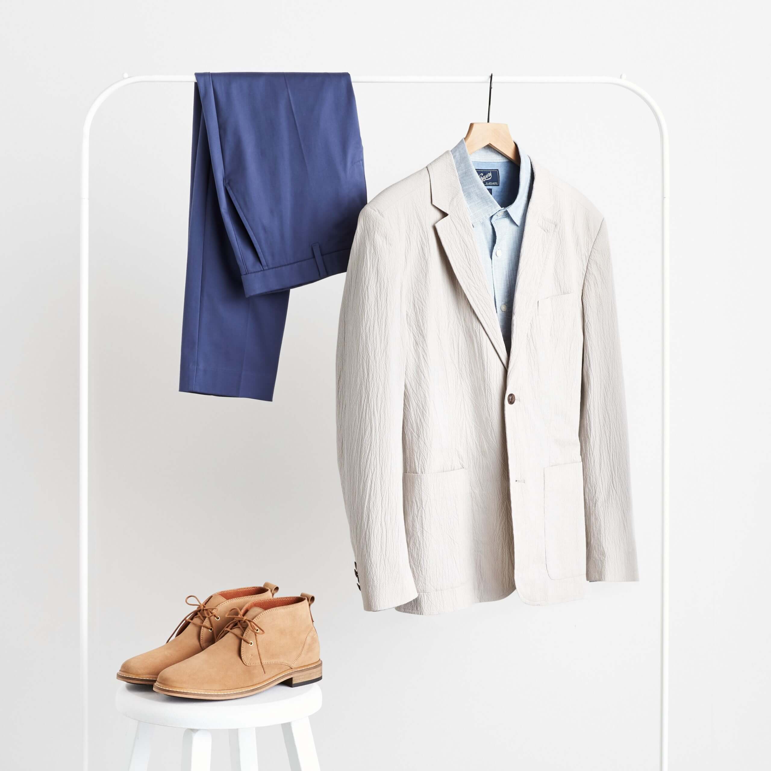 Stitch Fix beach wedding attire for men outfit featuring pants hanging over rack next to blazer over button-down shirt and chukka boots on stool.