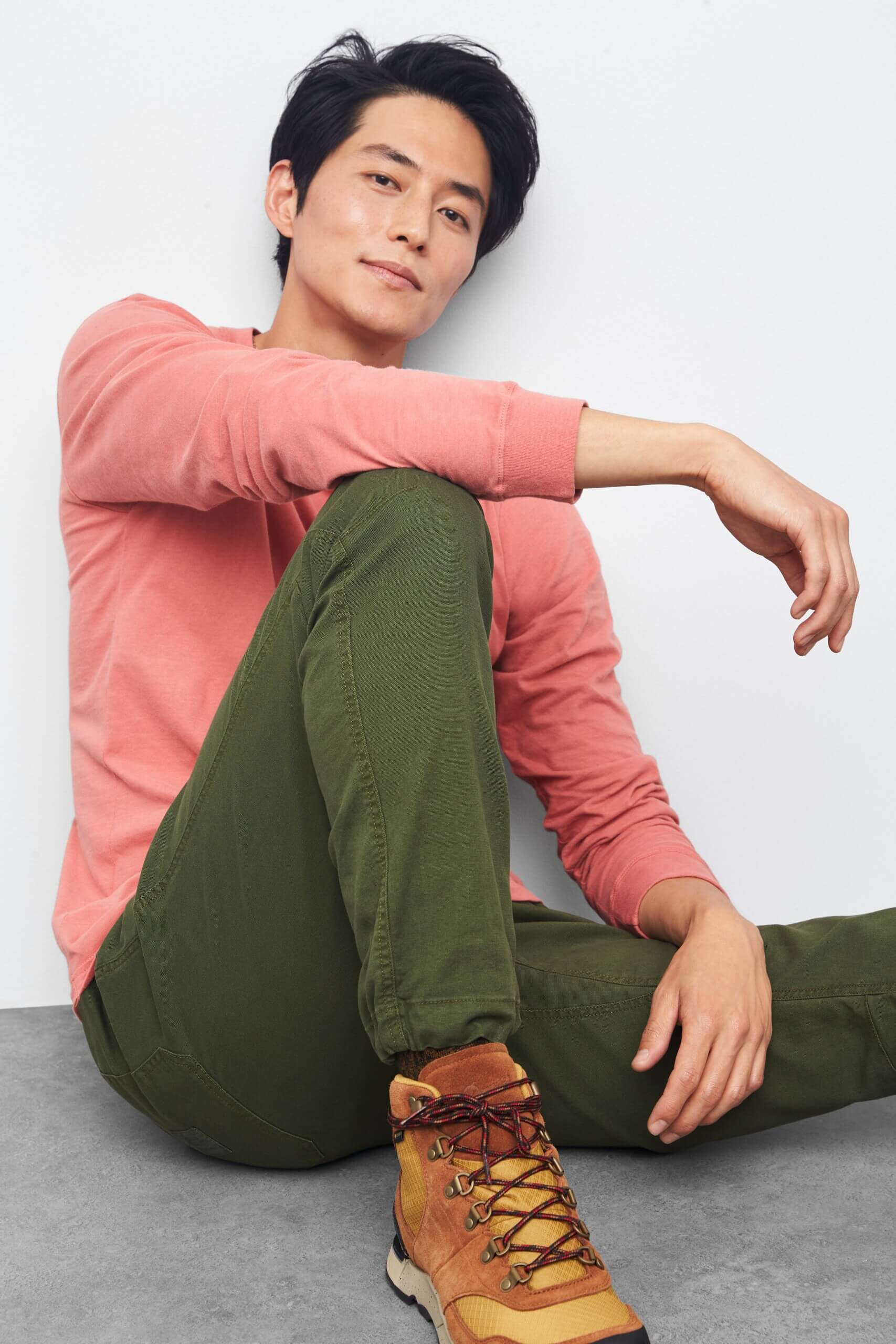 Stitch Fix model wearing men’s gorpcore outfit with salmon long-sleeved shirt, green pants, brown hiking boots, sitting down.