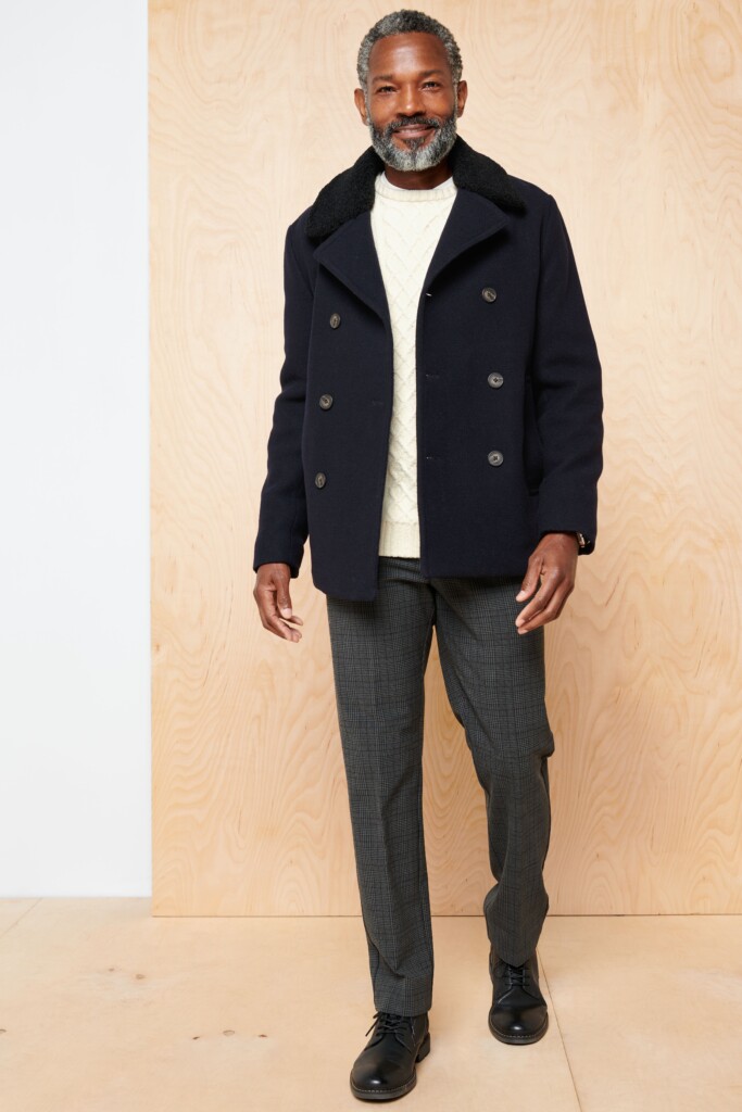 Stitch Fix model wearing preppy style men’s outfit featuring a black coat, white sweater, grey slacks, black leather shoes.