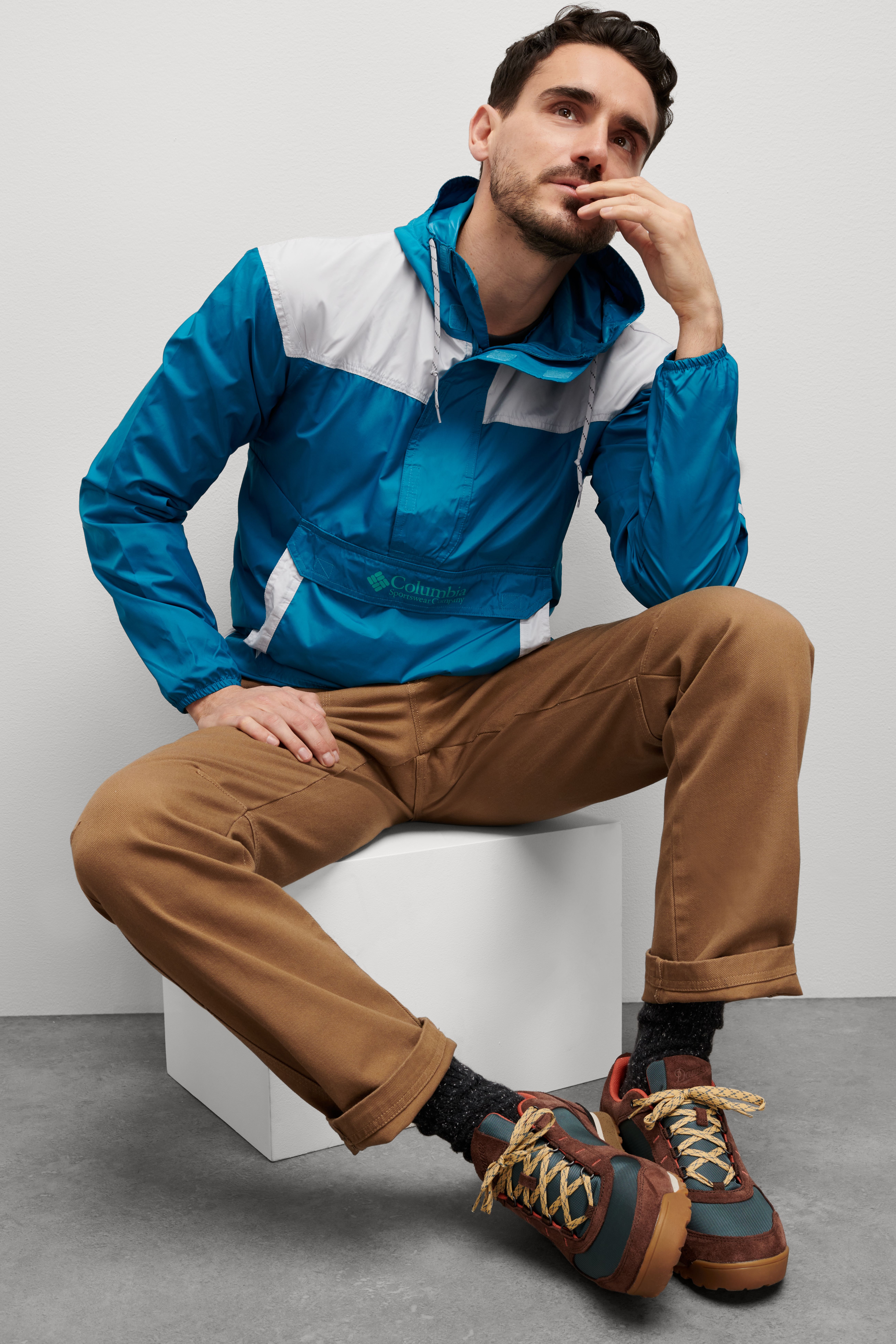 Stitch Fix model wearing men’s gorpcore outfit with blue and white jacket, tan pants and brown hiking boots, sitting down.