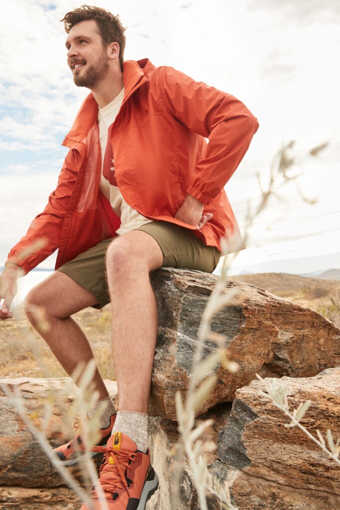Stitch Fix model wearing men’s 4th of July outfit with orange jacket, off-white T-shirt, olive shorts and orange sneakers.