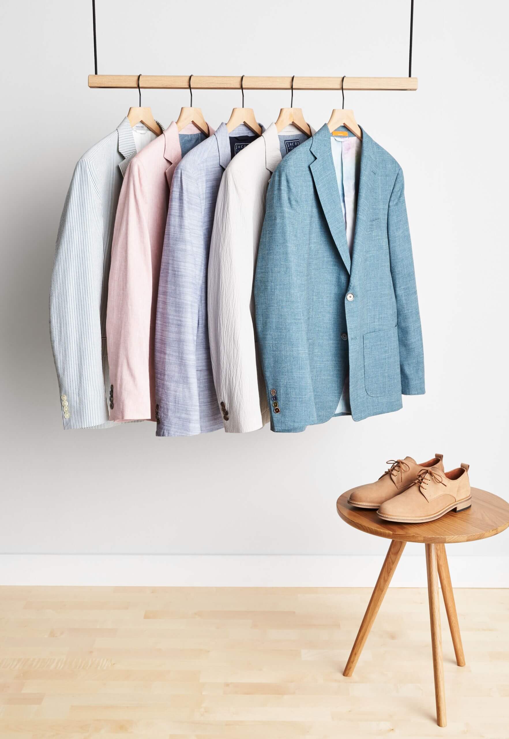Stitch Fix men’s spring wedding attire sport coats in various colors hanging on a rack next to brown oxford shoes on stool.
