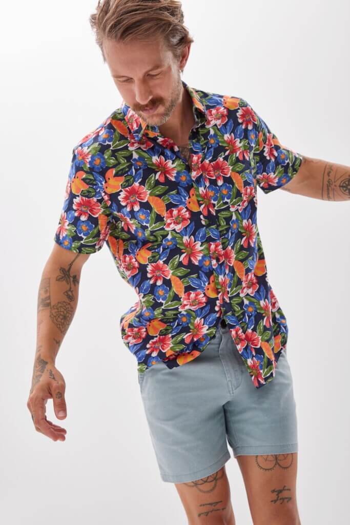 Man with tattoos stands and wears a floral summer shirt and denim shorts.