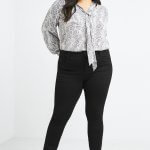 Stitch Fix women's model wearing plus-size clothing featuring snakeskin print tie-neck blouse, black skinny pants and black heels.