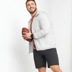Stitch Fix model wearing a red T-shirt, grey zip-up hoodie, black shorts and black watch holding a football.