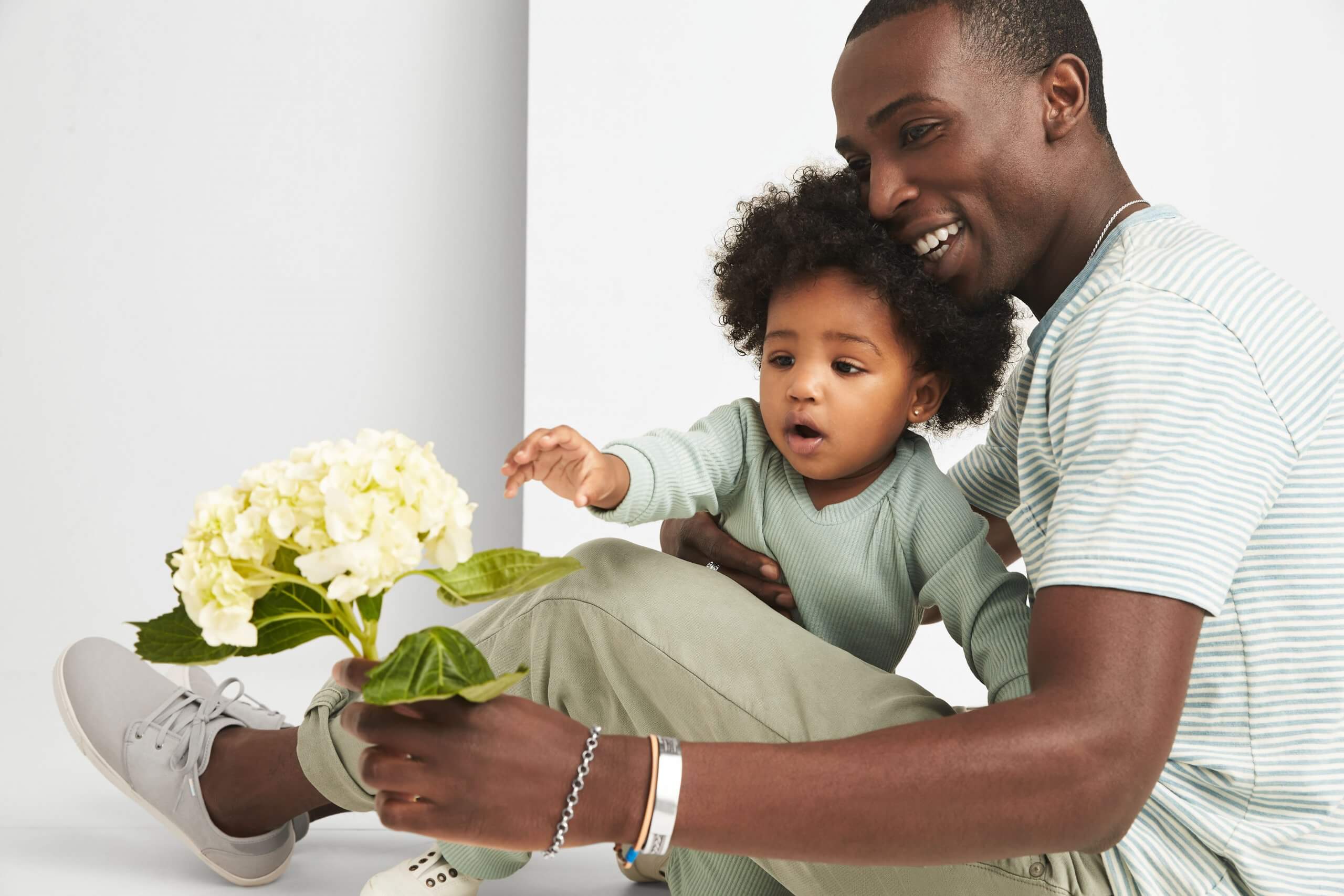 Stitch Fix Model wearing green shirt, khaki pants, grey sneakers and silver bracelets holding a cream flower and a child wearing a green shirt.