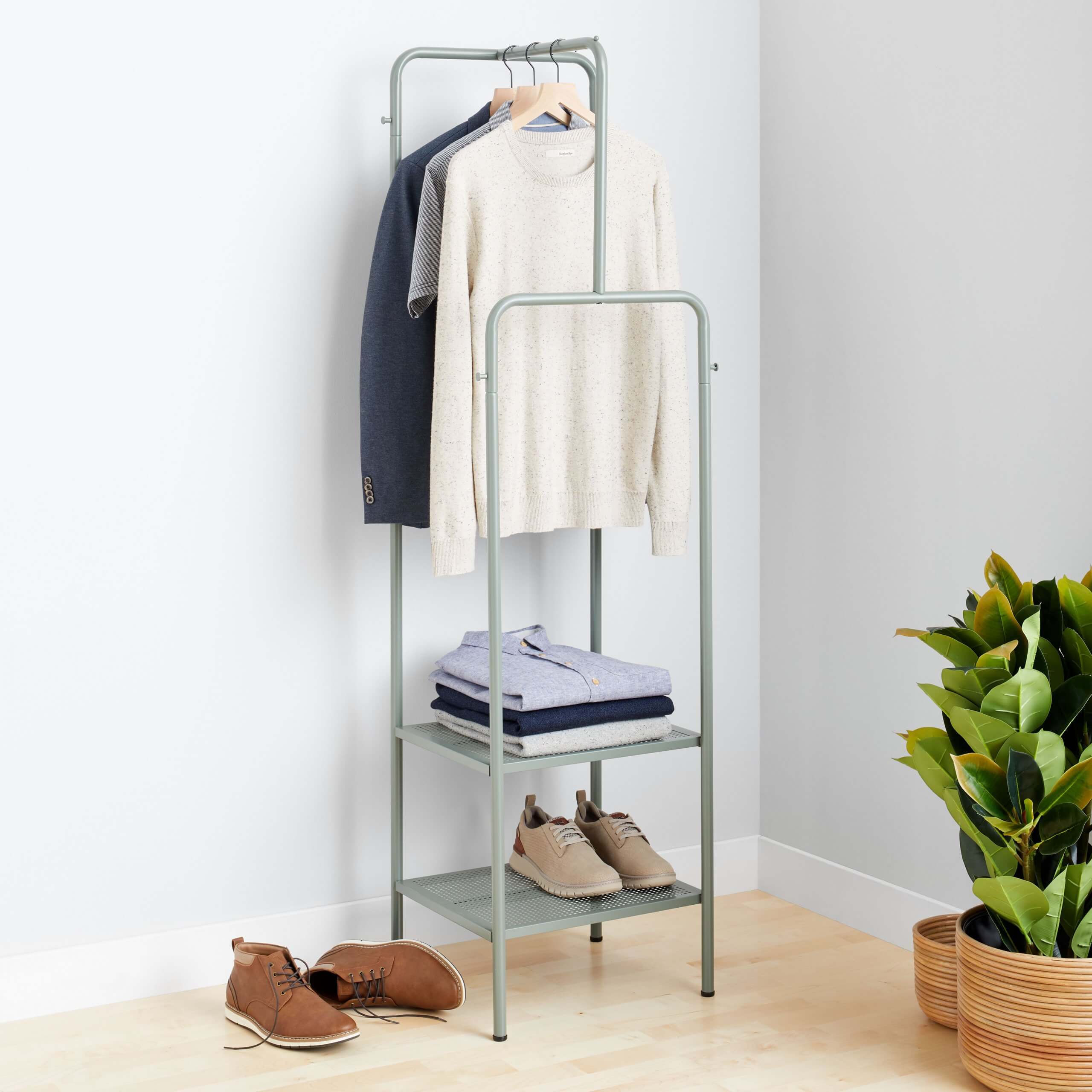 Stitch Fix Men’s brown chukkas next to clothing rack with blue blazer, grey T-shirt and off-white pullover hanging above stack of button-downs and beige shoes. Two potted plants in the bottom right corner.