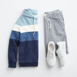 Stitch Fix Men’s outfit laydown featuring a blue, navy and white colorblock hoodie, grey shorts and white sneakers.