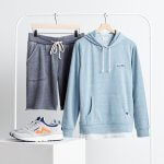 Stitch Fix Men’s outfit featuring navy French terry shorts and light blue hoodie hanging on a clothing rack with grey sneakers with orange detailing on a table.