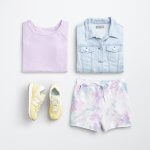 Stitch Fix women’s jean jacket outfit laydown with a light purple crew neck sweatshirt, light blue denim jacket, yellow sneakers and purple, white and blue tie-dye drawstring shorts.