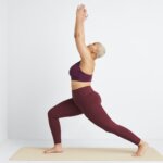 Stitch Fix women's model in warrior 1 pose wearing a yoga outfit featuring matching burgundy sports bra and leggings.