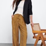 Stitch Fix model wearing women's 90s fashion outfit with white shirt, navy blazer, gold jewelry, satin pants and loafers leaning on chair.