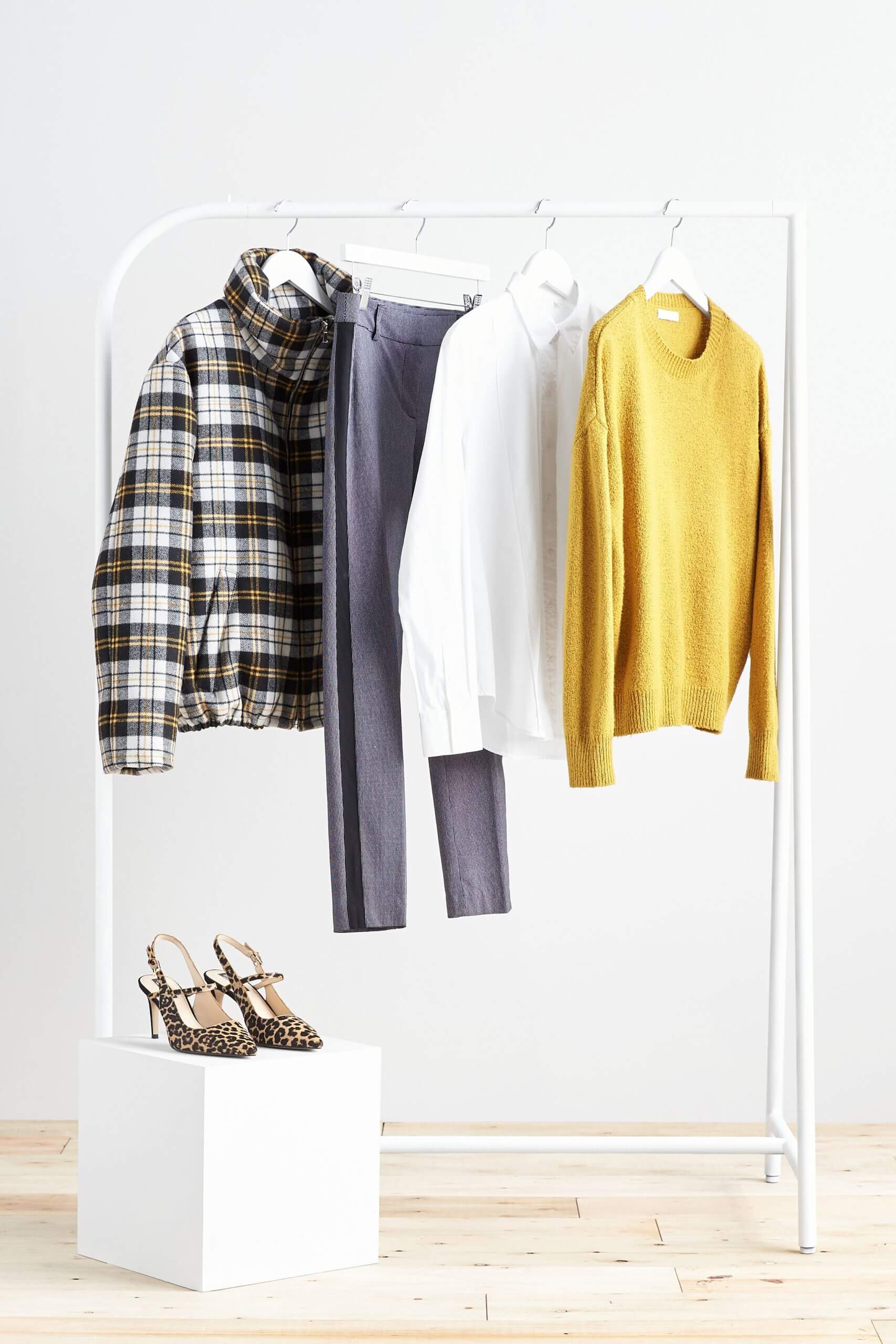 Stitch Fix Women's rack image featuring yellow sweater, white button-down shirt, blue pants and plaid puffer jacket hanging on white rack, next to animal-print heels. 