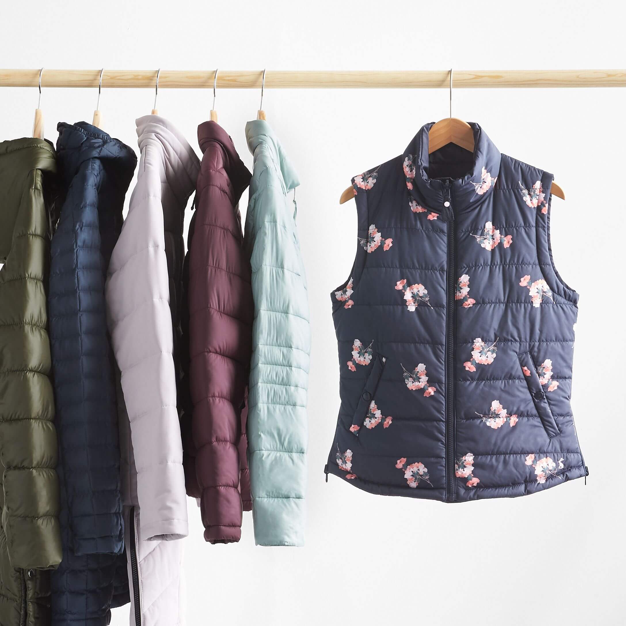 Stitch Fix Women's rack image featuring navy vest with floral print and puffer jackets in light blue, burgundy, lilac, navy and olive green tones hanging on wooden pole. 