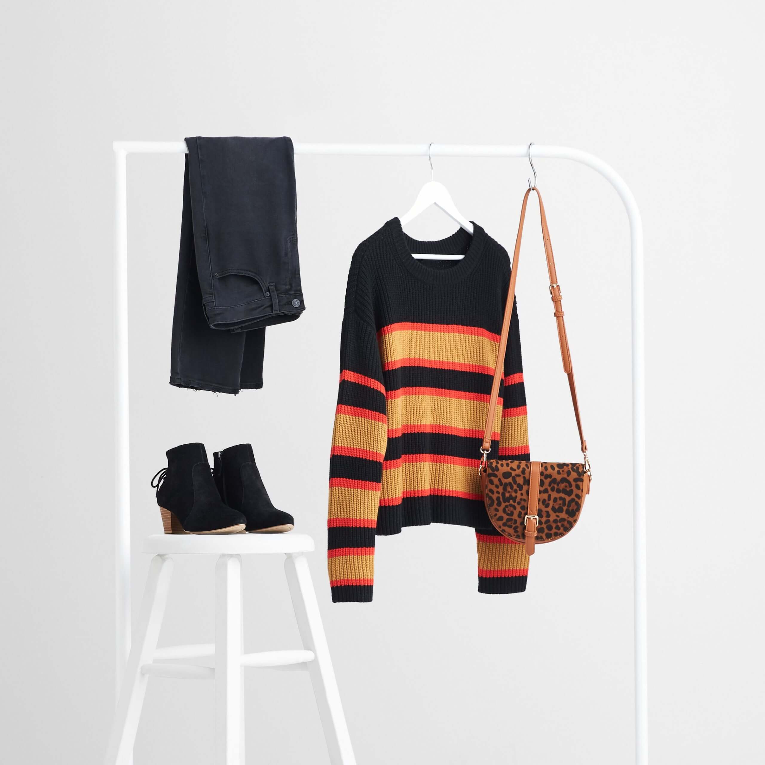 Stitch Fix Women's rack image with brown animal-print purse, black pullover sweater with orange stripes and black jeans on white rack, next to black booties on a white stool.
