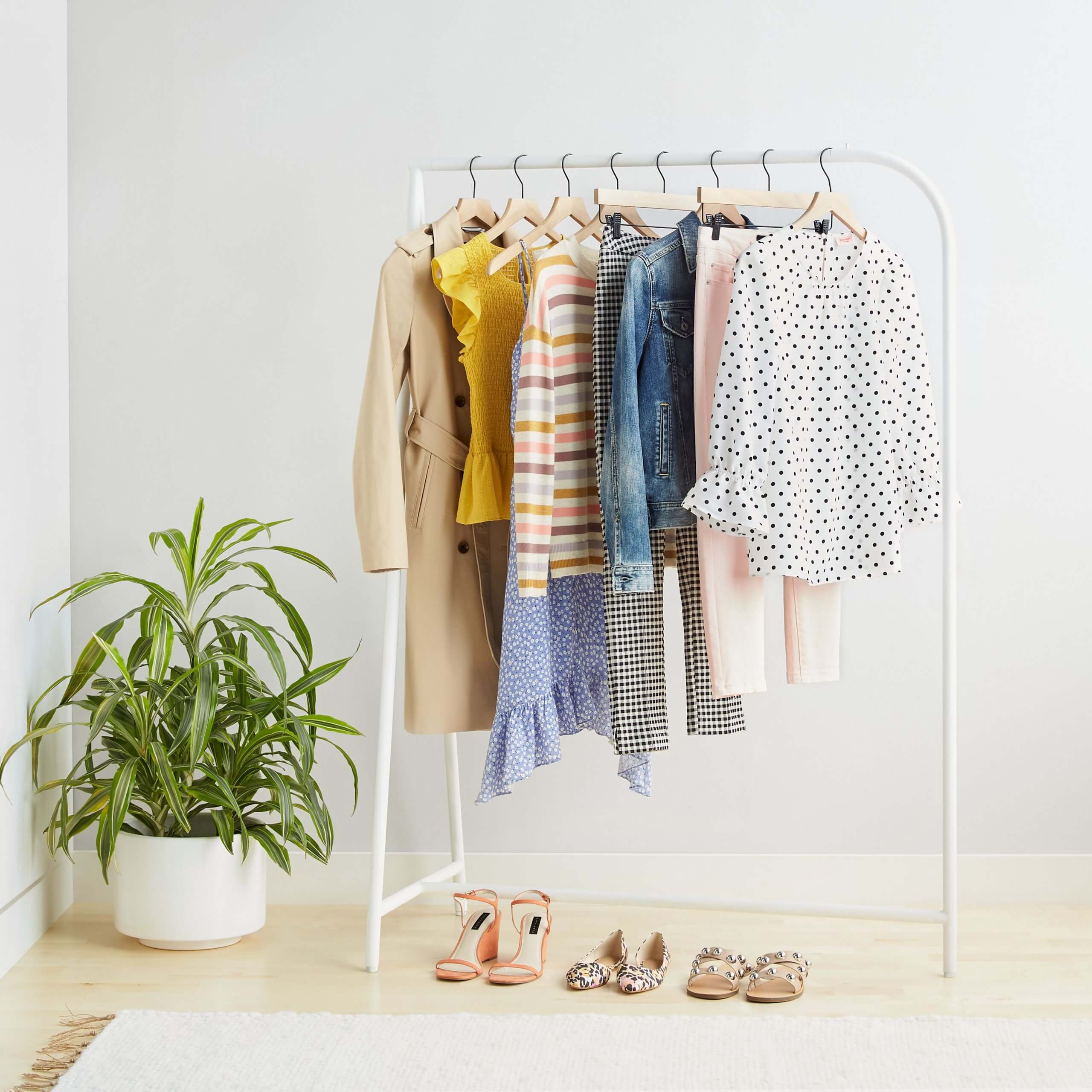Stitch Fix Women’s rack image featuring white polka dot blouse, pink jeans, blue denim jacket, multi-colored striped pullover, yellow blouse and tan trench hanging on white rack with tan slide sandals, animal-print flats and coral wedges on the ground. 