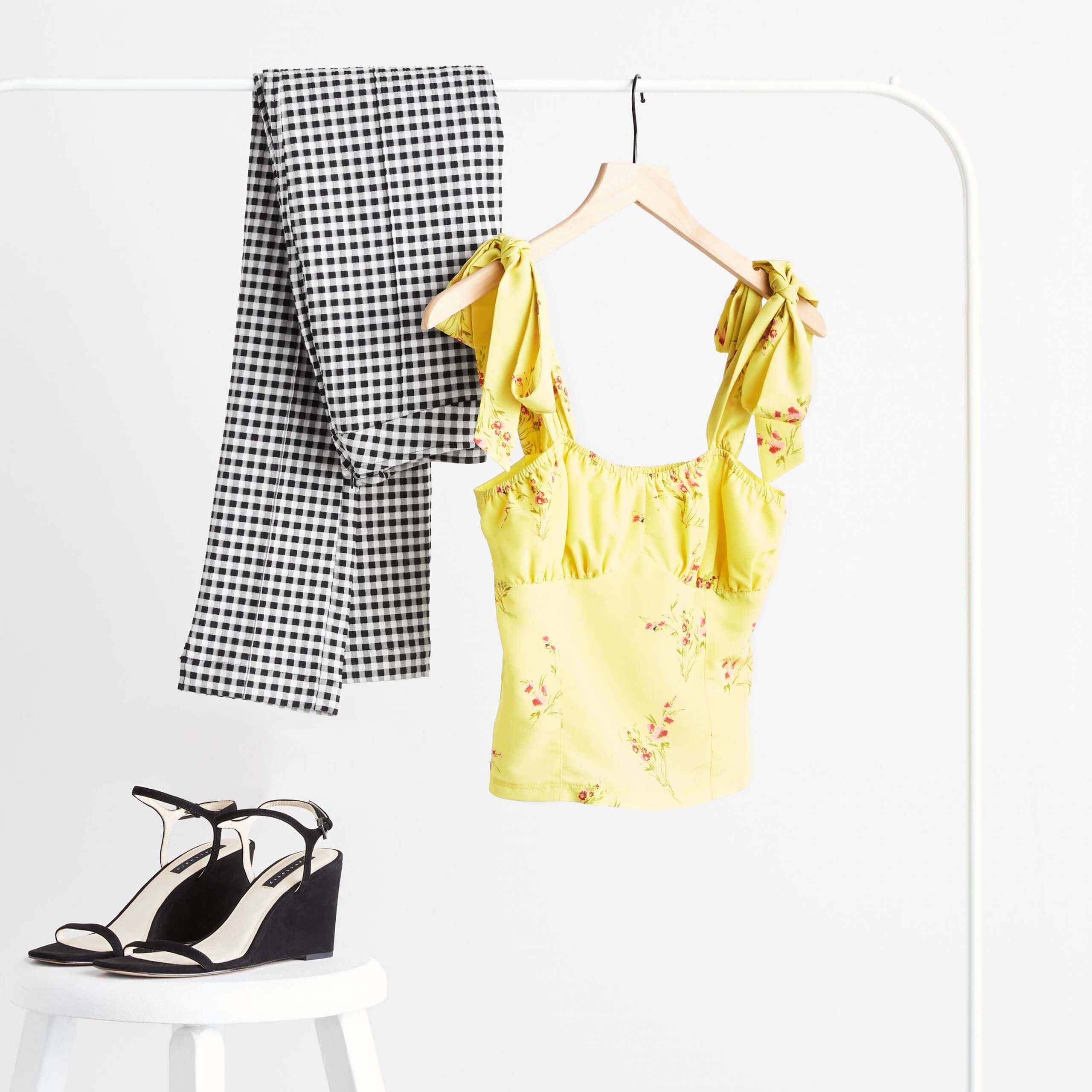 Stitch Fix Women's yellow floral ruffle sleeve top and black and white checkered pants hanging on rack next to black wedges on white stool. 