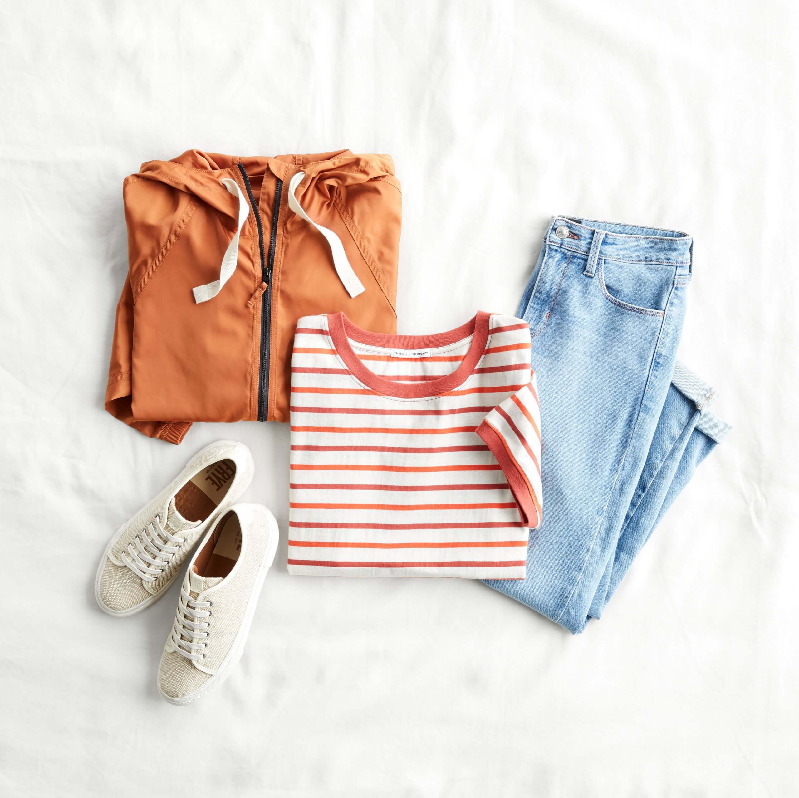 Stitch Fix Women's outfit laydown featuring orange and white striped tee, lightwash jeans, white sneakers and orange rain jacket. 