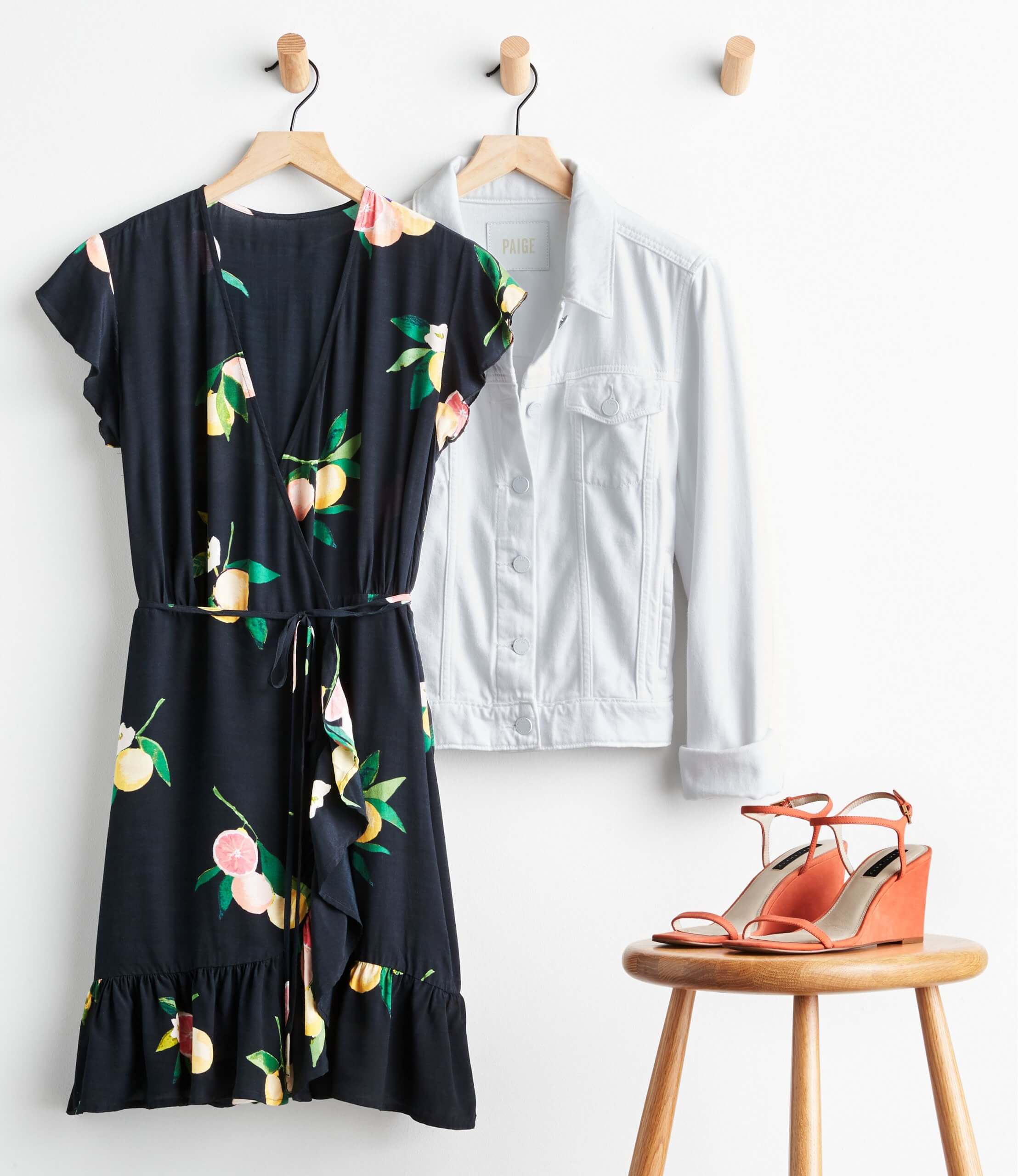 Stitch Fix women’s outfit featuring black floral print wrap dress with tie-waist, ruffle hem with white denim jacket on hanger and coral wedge heels on stool.