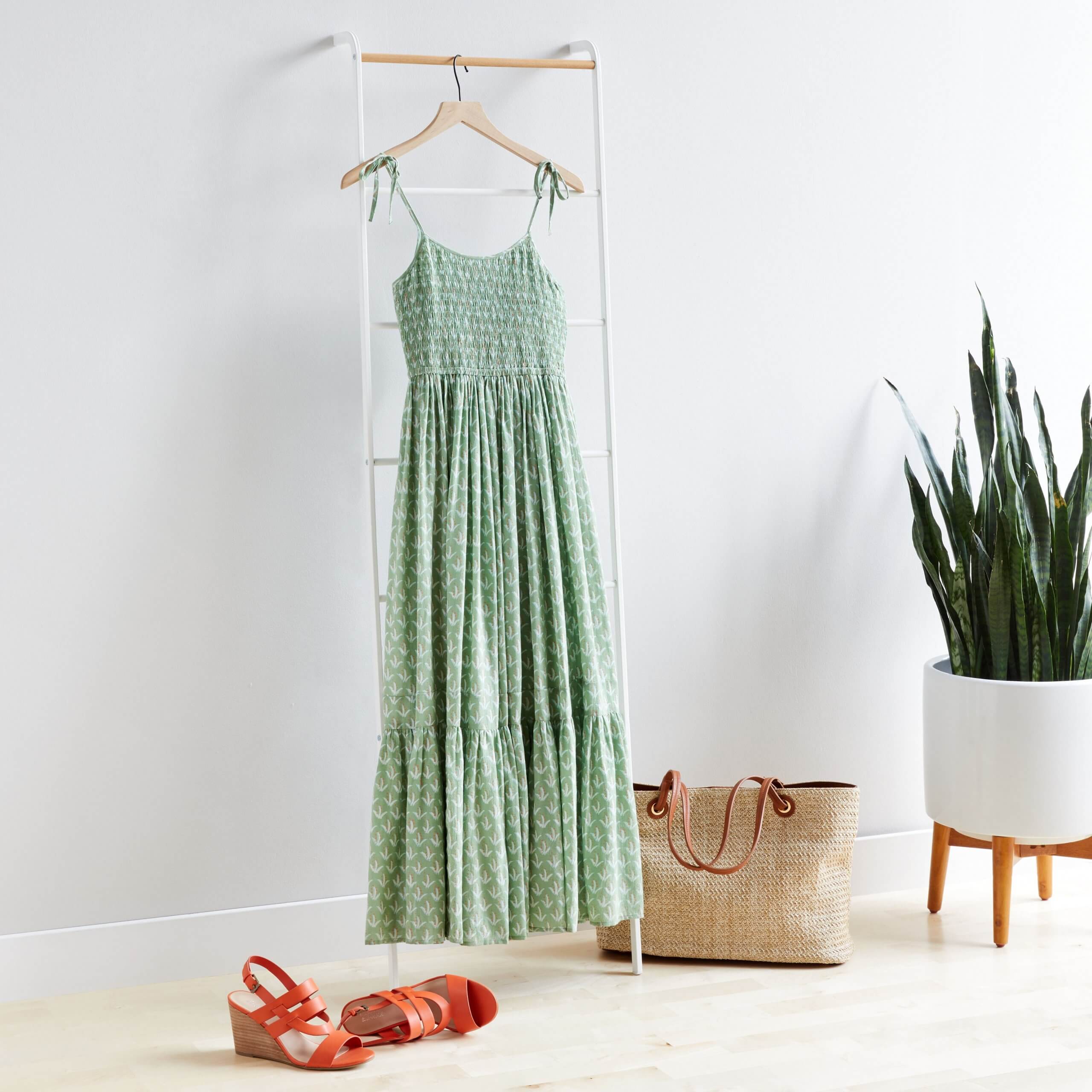 Stitch Fix Women’s green maxi dress hanging on white rack next to tan tote bag and orange wedges. 
