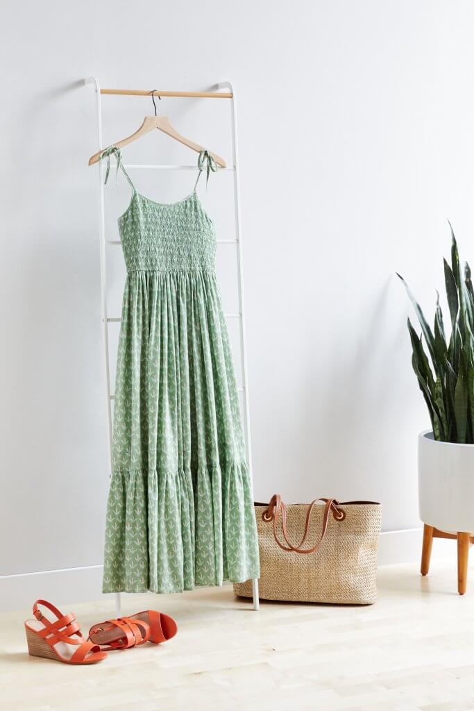 Stitch Fix Women’s green tiered maxi dress hanging on clothing rack next to a potted plant. Featured with straw tote and red wedge sandals.