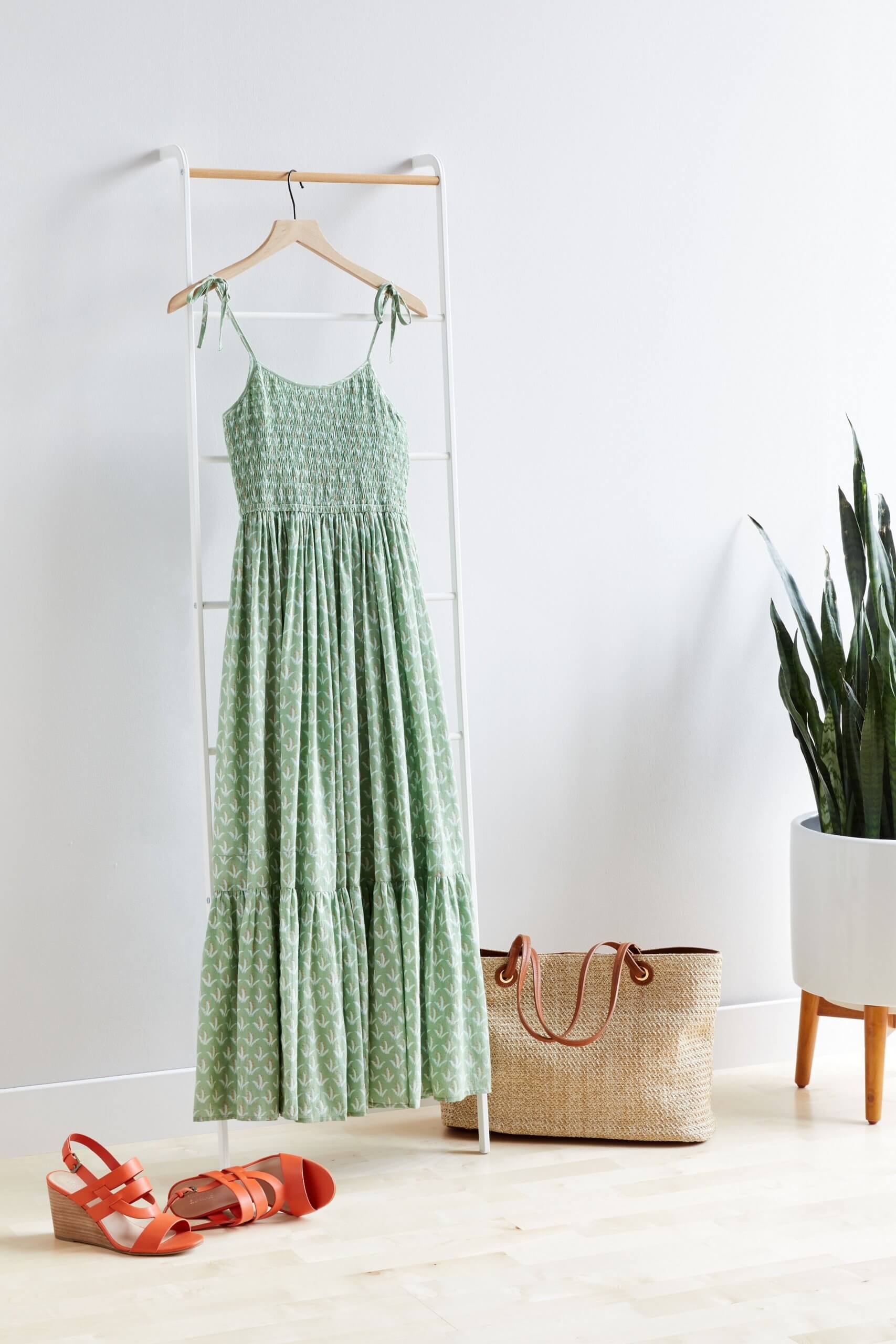 Stitch Fix Women’s green tiered maxi dress hanging on clothing rack next to a potted plant. Featured with straw tote and red wedge sandals.