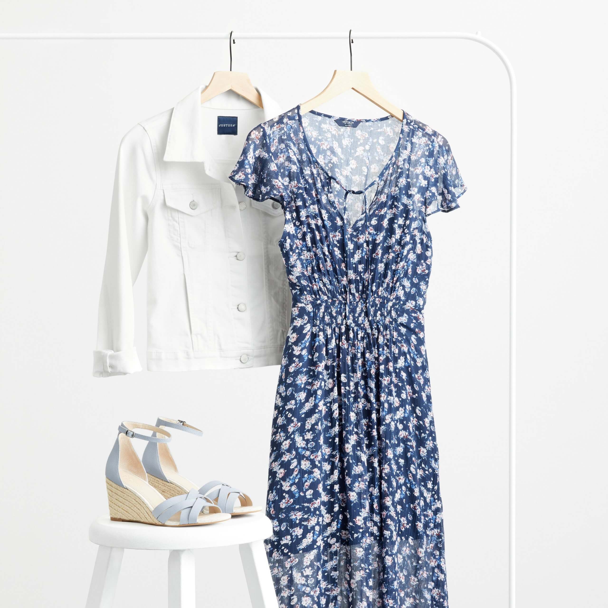 Stitch Fix Women’s rack image featuring white denim jacket and blue and white printed midi dress on hangers on white rack, next to blue wedges on a white stool.