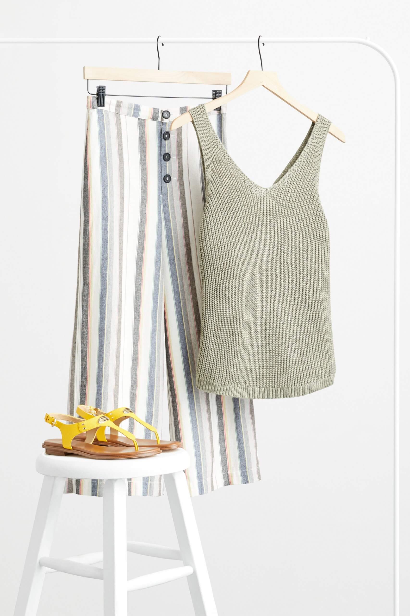 Stitch Fix Women’s olive v-neck sweater tank and striped wide-leg pants hanging on white rack next to yellow sandals on a white stool. 