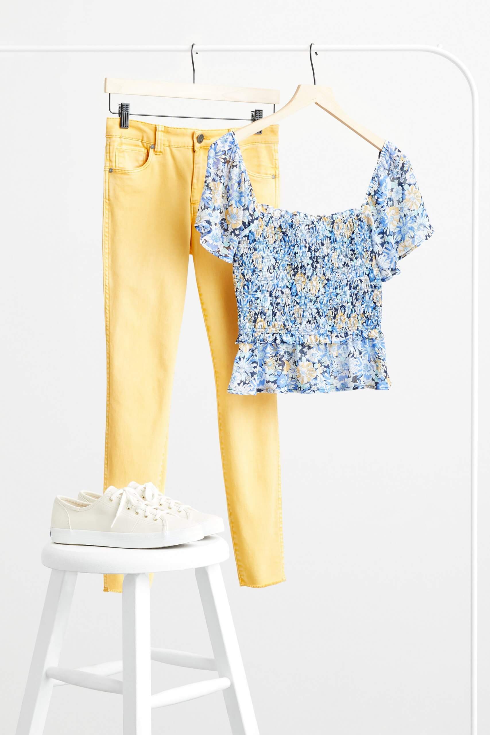 Stitch Fix women’s yellow skinny jeans and blue floral blouse hanging on rack. White sneakers on a white stool.