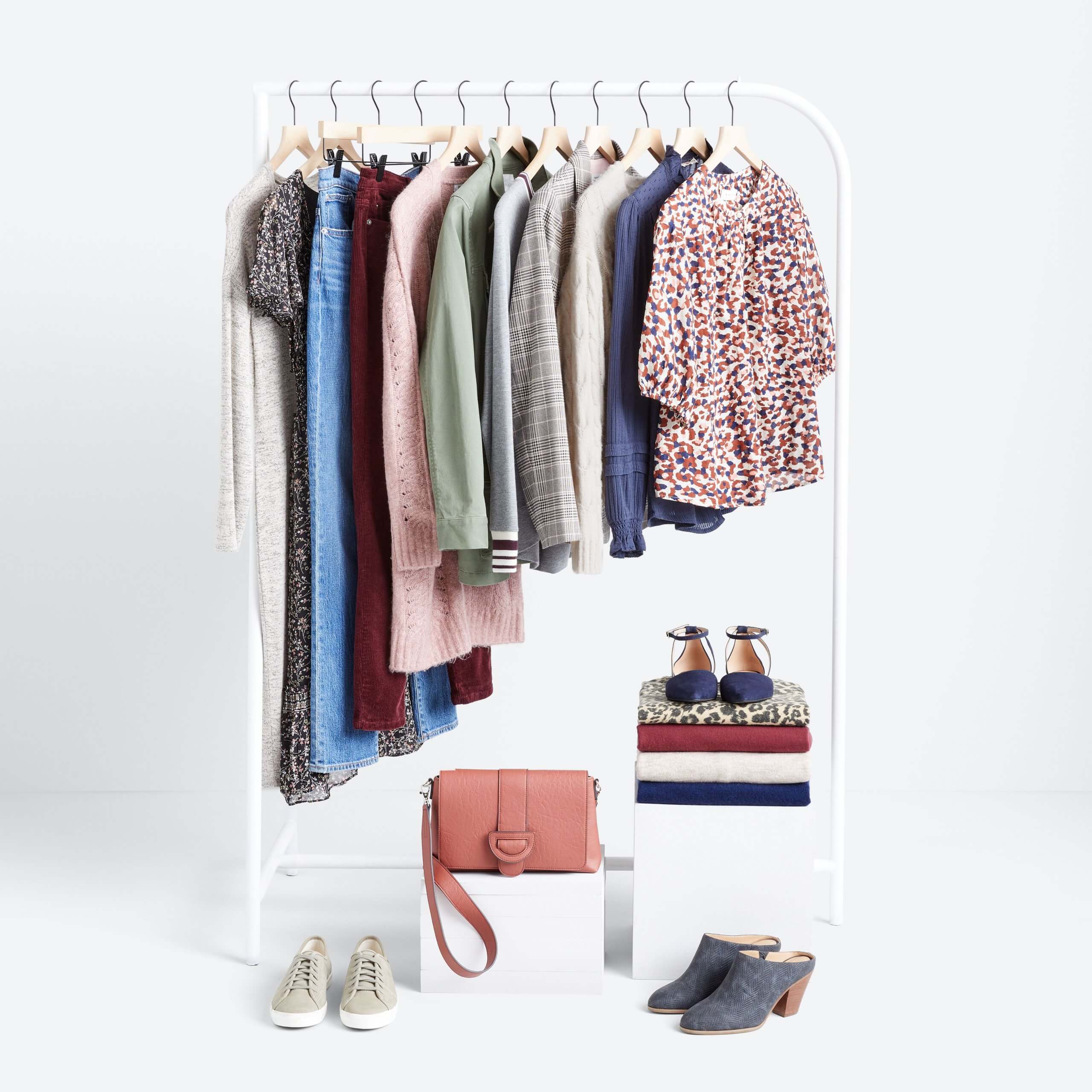 Stitch Fix Women’s rack image featuring red, wnite and blue printed blouse, navy blouse, cream pullover, plaid blazer, grey pullover, green jacket, pink cardigan, burgundy jeans, blue jeans and black printed dress hanging on white rack with tan sneakers and navy mules on the floor, next to pink bag and folded shirts in navy, tan and burgundy, with animal-print flats on white block. 