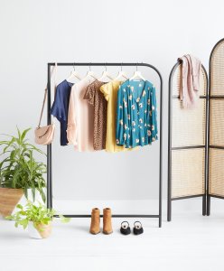Stitch Fix women’s beige purse on a rack with five tops in an assortment of colors and patterns next to two potted plants, boots and a pair of flats. A room divider with a blush cardigan slung over it.