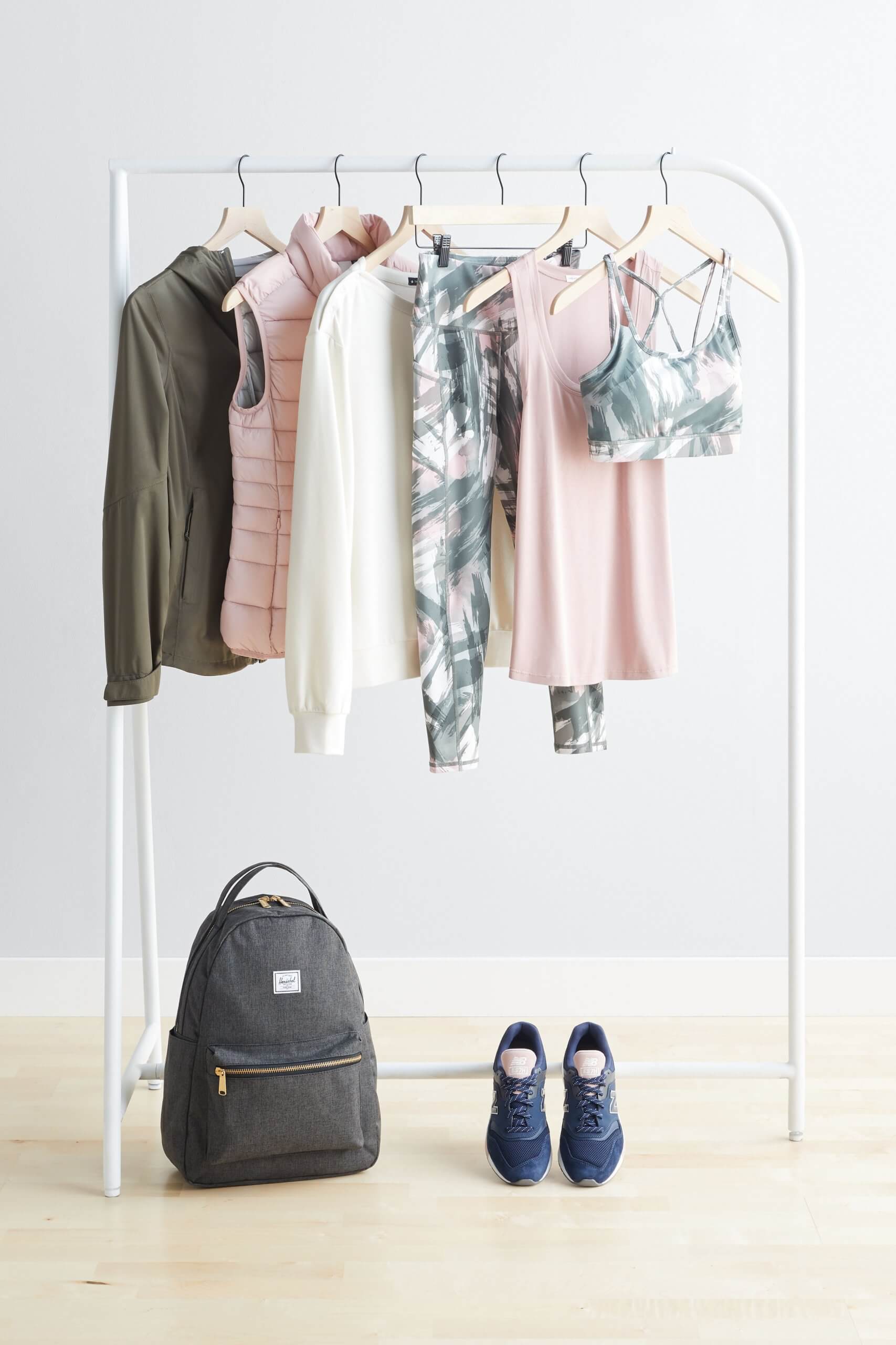 Stitch Fix Women’s olive jacket, pink puffer vest, white crew neck sweatshirt, tropical print leggings, pink tank and tropical print sports bra on a rack. Grey backpack and navy sneakers on the floor.