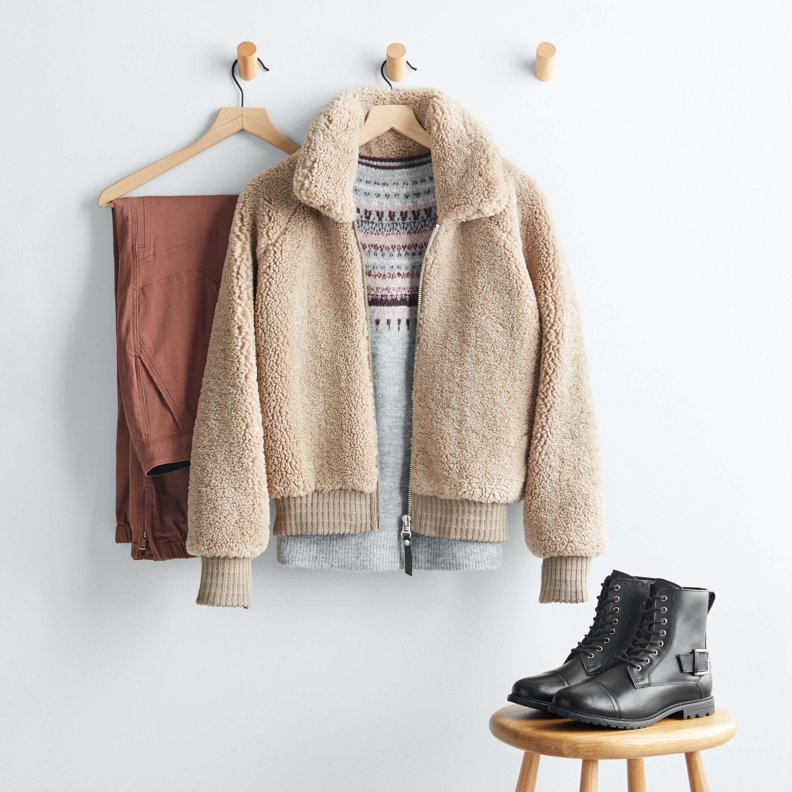 Stitch Fix women’s cabincore outfit with brown pants next to blue Fair Isle sweater under tan sherpa jacket hanging up next to black boots on stool.