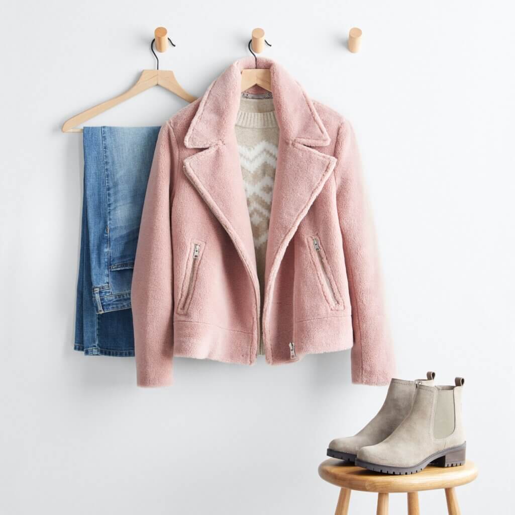 Stitch Fix women’s winter fashion 2021 outfit with light-wash denim, tan intarsia sweater and pink sherpa jacket on hangers with taupe Chelsea boots on stool.