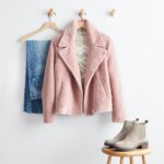 Stitch Fix women’s winter fashion 2021 outfit with light-wash denim, tan intarsia sweater and pink sherpa jacket on hangers with taupe Chelsea boots on stool.