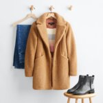 Stitch Fix women’s boots with jeans outfit featuring dark wash jeans, multi-colored striped sweater under a tan wubby coat on hangers and black Chelsea boots on a stool.