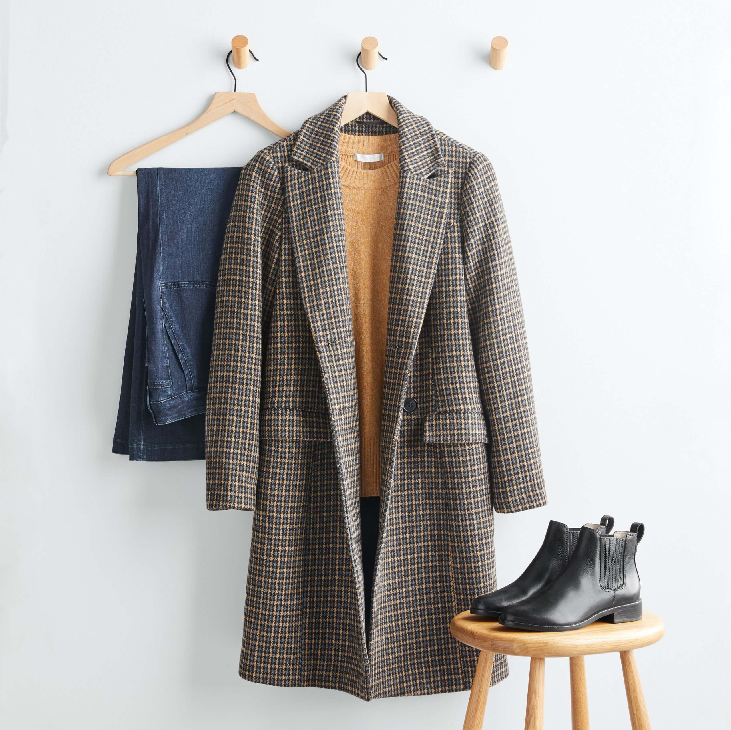 Stitch Fix Women’s outfit featuring brown plaid coat over mustard sweater hanging on wooden hanger, dark wash jeans on wooden hanger next to black leather booties on wooden stool. 