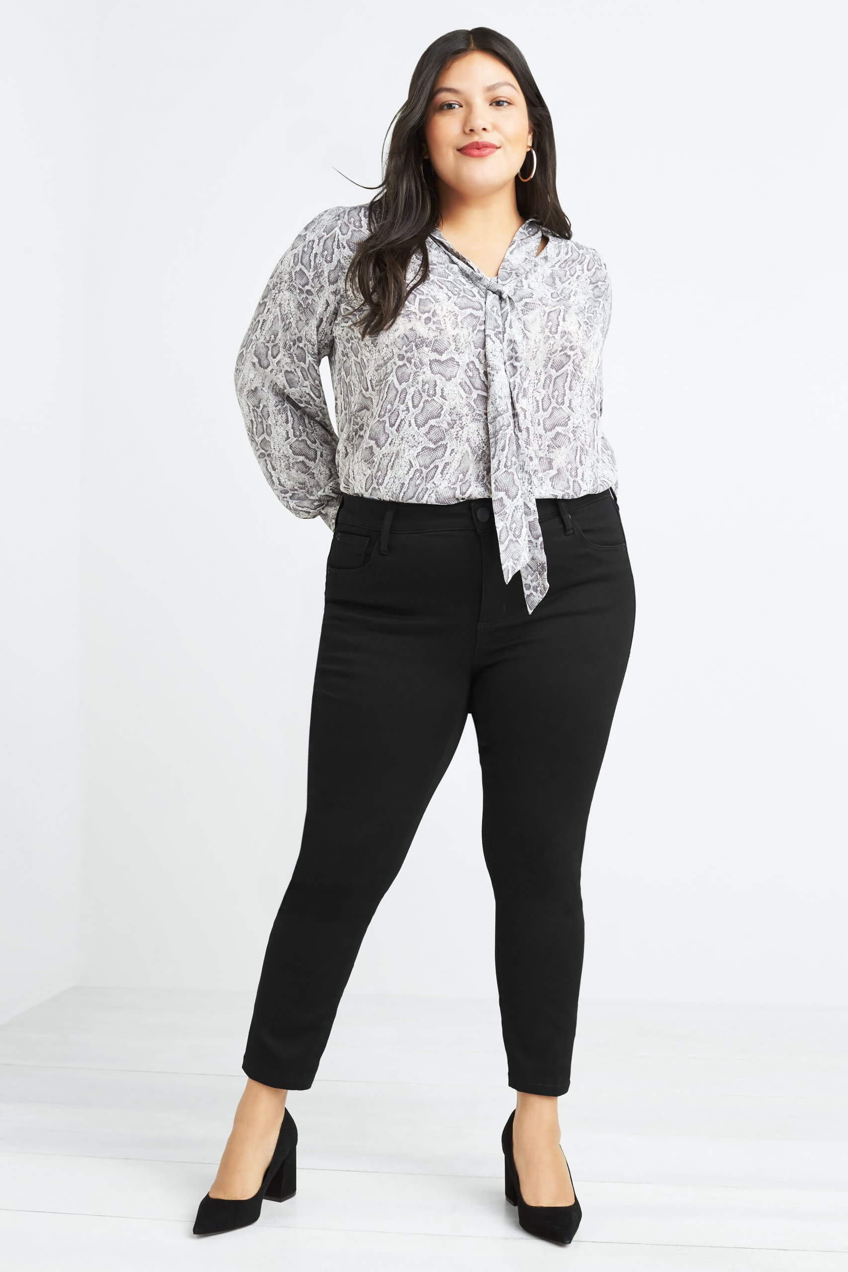 Stitch Fix women's model wearing plus-size clothing featuring snakeskin print tie-neck blouse, black skinny pants and black heels.