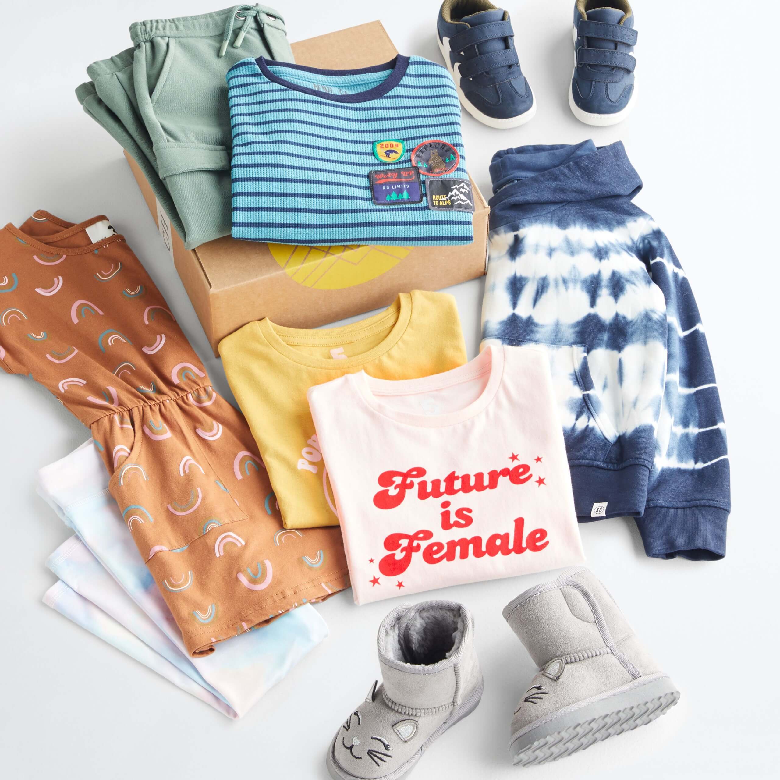 Stitch Fix Kid's outfit laydown featuring green pants and blue striped shirt on Stitch Fix delivery box, next to navy blue sneakers, navy blue tie-dye hoodie, pink and yellow t-shirts, silver boots, brown dress with rainbow print and light blue and light pink tie-dye leggings. 