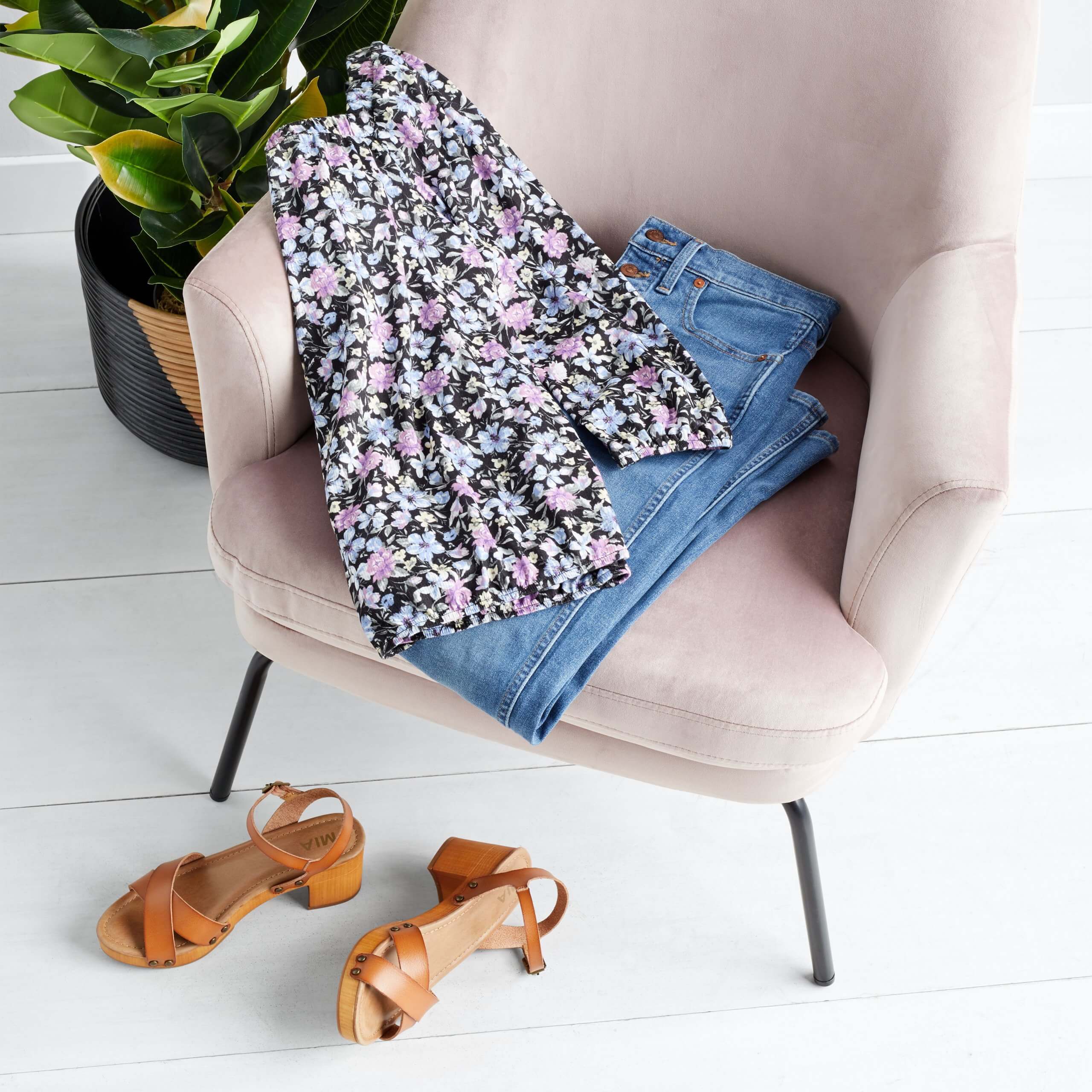 Stitch Fix women’s blue jeans and floral blouse on a pink chair next to potted plant and leather heel sandals on floor.