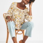 Stitch Fix sitting model in women’s floral print blouse, light-wash jeans and brown sandals with ankle straps.