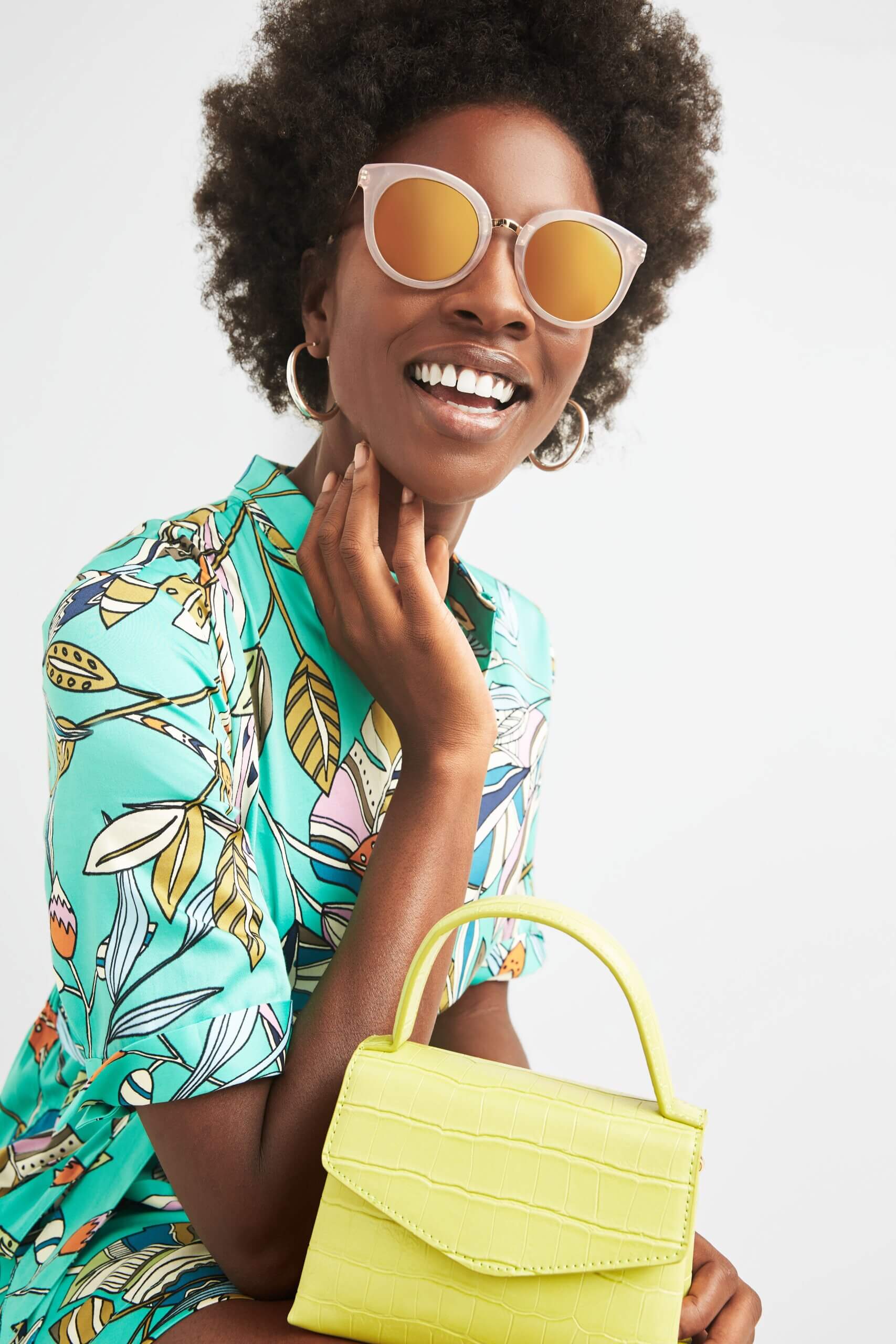 Stitch Fix Women's model wearing gold hoop earrings, sunglasses, green leaf-print romper and a lime green clutch.