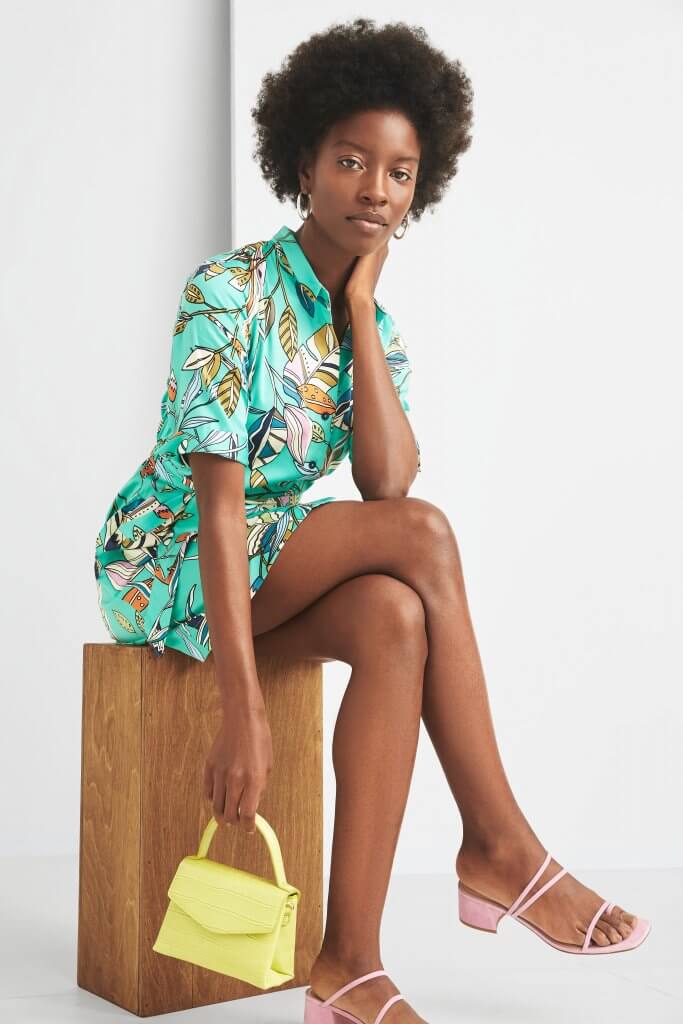 Stitch Fix model wearing gold hoop earrings, a green, leaf-printed romper, pink strappy heels and a lime green mini purse.
