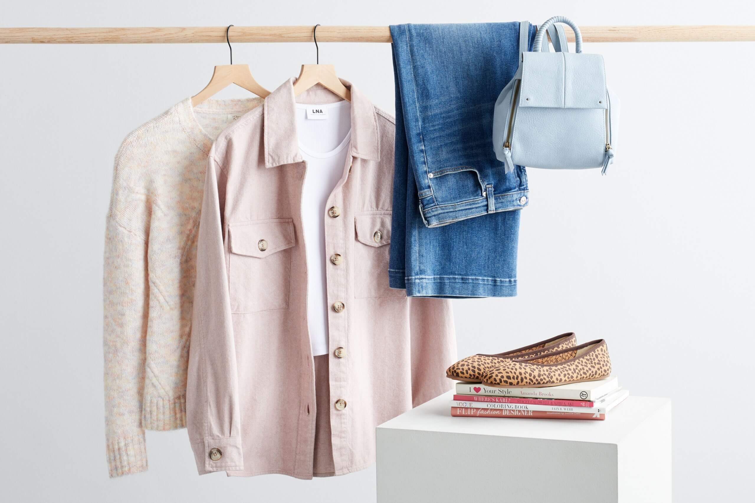 Stitch Fix women's shacket outfit featuring a white sweater, pink shacket over white T-shirt, blue jeans and light blue backpack on rack next to animal print flats on a stack of books.