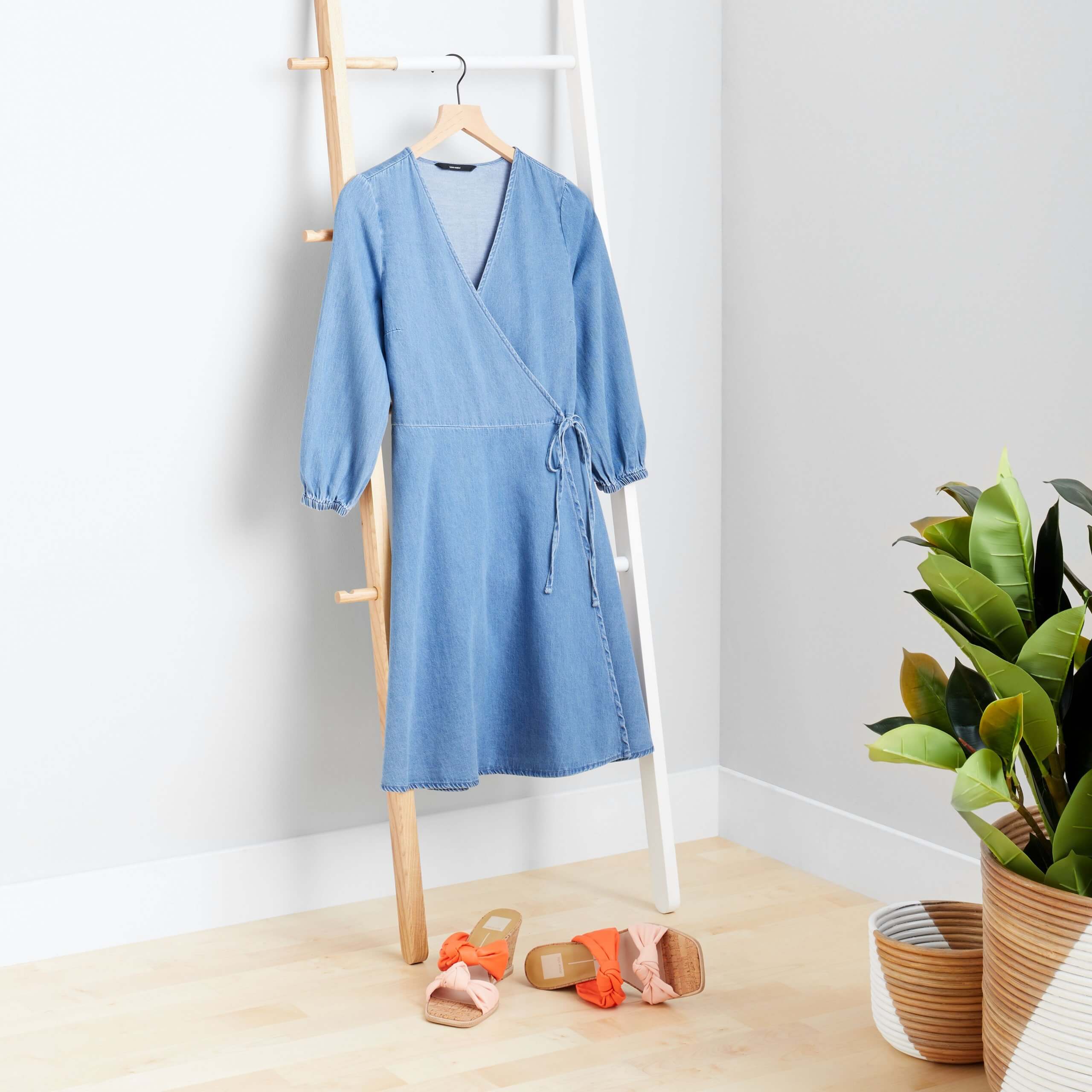 Stitch Fix Women's blue wrap dress hanging on wooden ladder next to orange and pink slide wedges. 