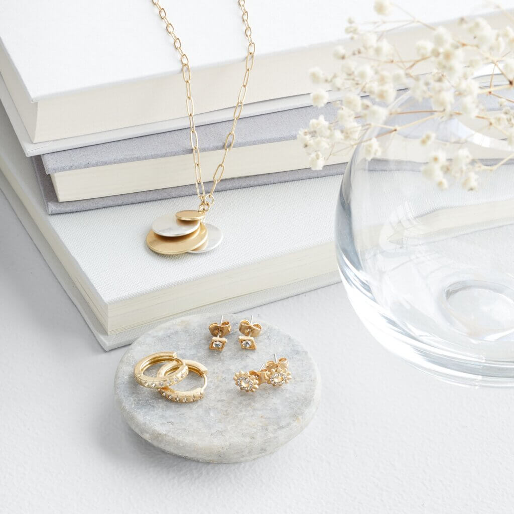 Stitch Fix women’s gold earrings on a dish and mixed-metal necklace laying on a stack of books next to a vase with flowers.