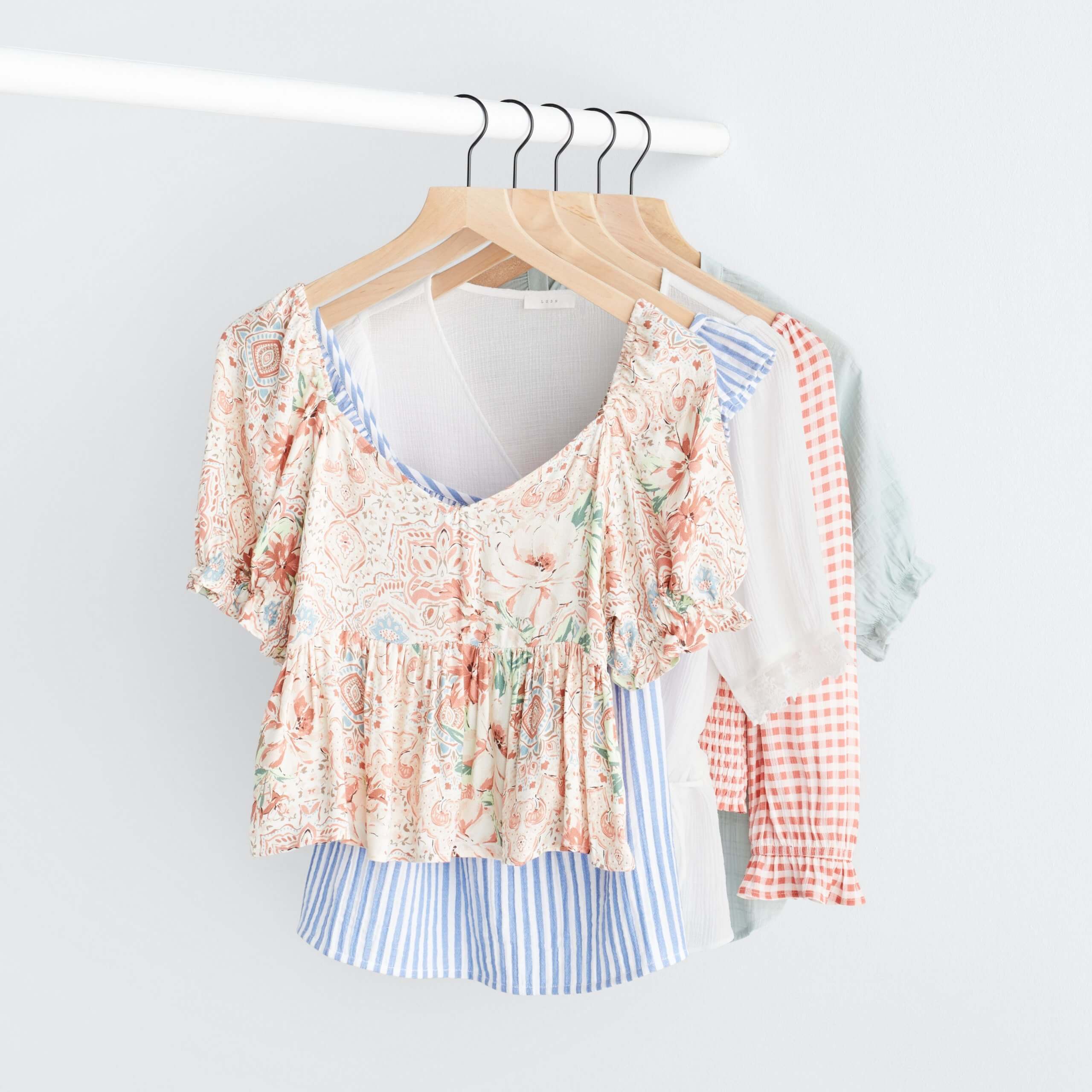 Stitch Fix Women’s rack image with white peplum top with pink florals, blue and white striped ruffle tank, white peplum top, pink and white gingham top and green ruffle sleeve top hanging on white pole.