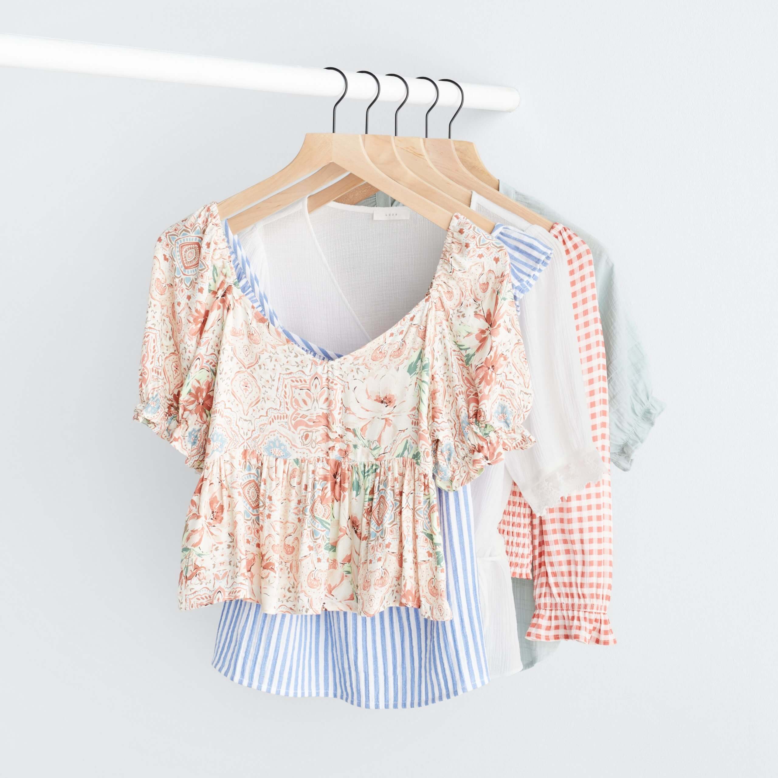 Stitch Fix Women’s rack image with white peplum top with pink florals, blue and white striped ruffle tank, white peplum top, pink and white gingham top and green ruffle sleeve top hanging on white pole. 